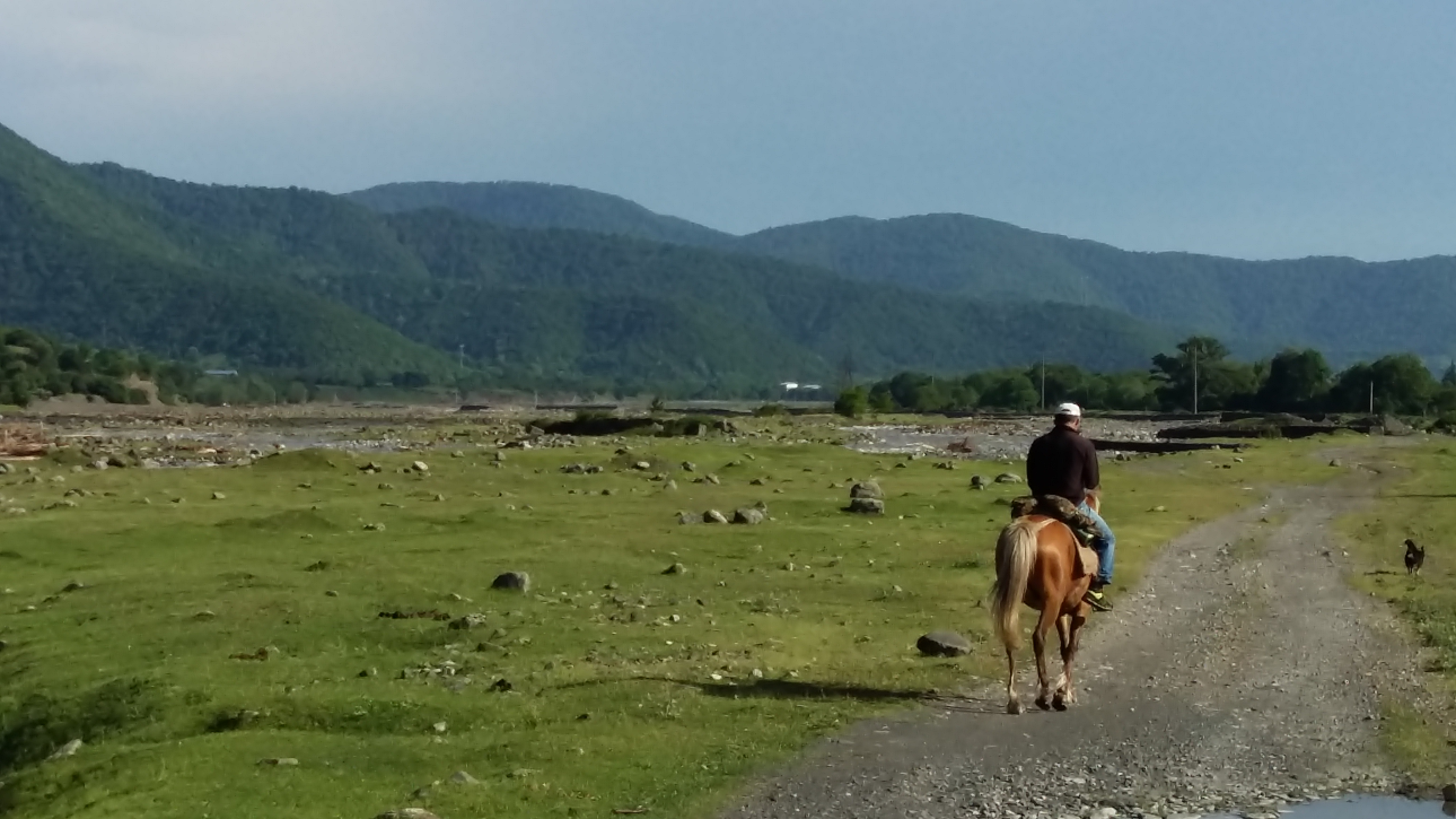 Image of a horseman on a trail through a wide river meadow full of stones, with mountains in the background.