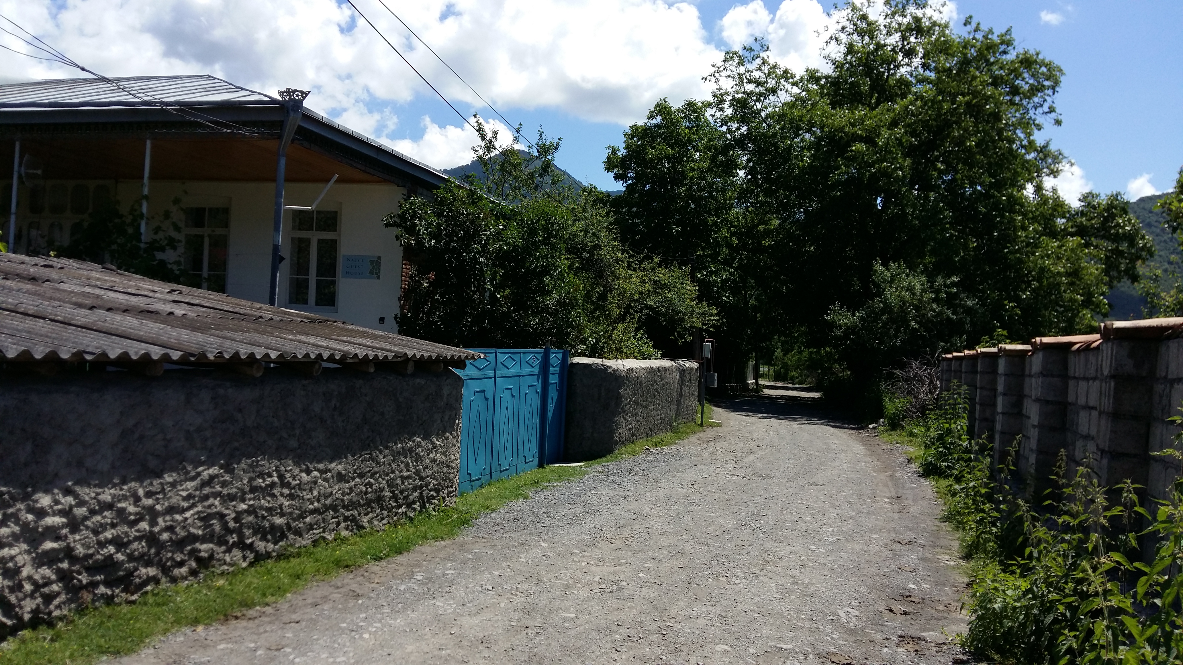 A dirt road with walls on either side, one of which has a metal gate. Behind the gate, a house is visible.