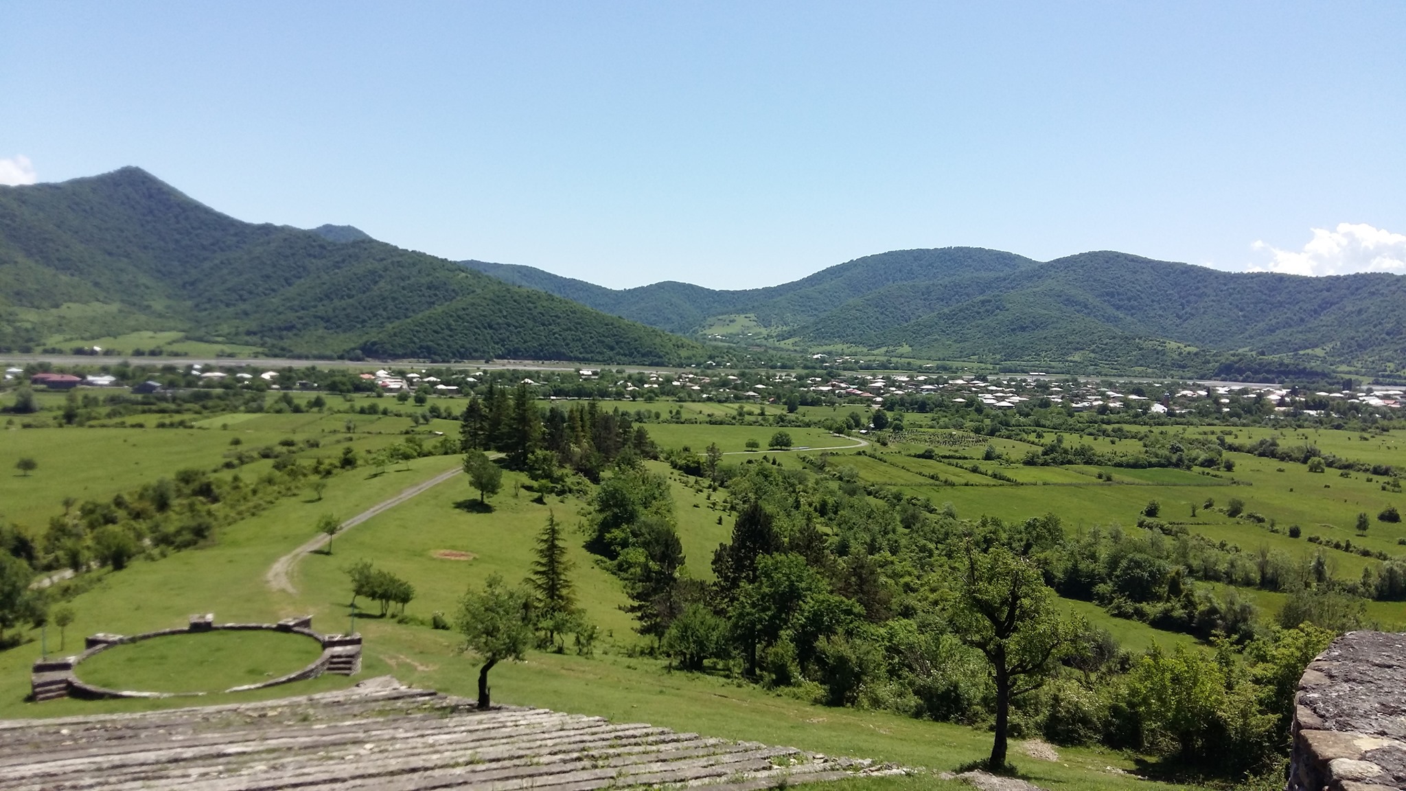 View of pastures, with a village and forested mountains in the distance.