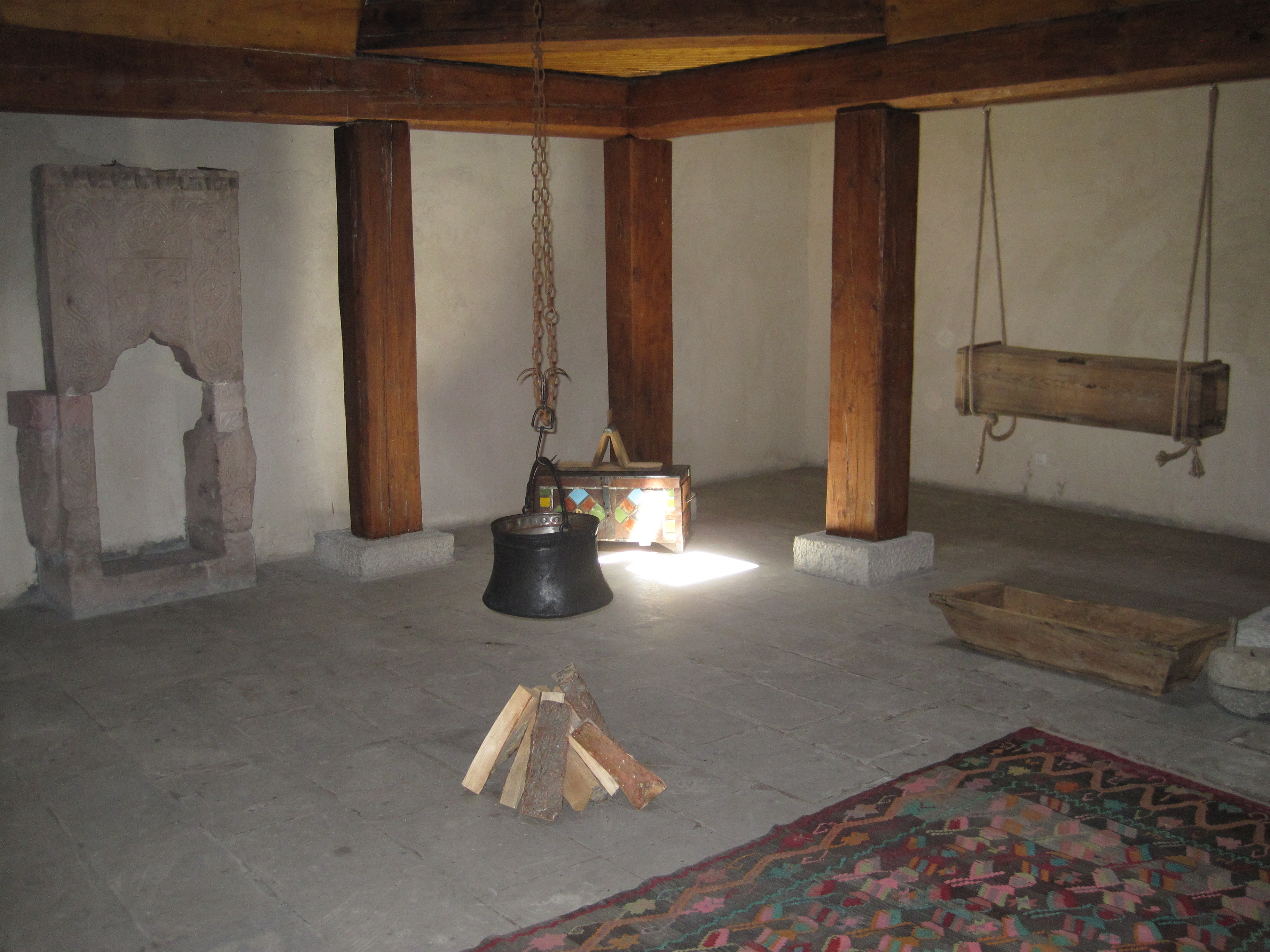 Interior with central hearth, kettle on hearth chain, chest with wool carding comb, corn crib (?), kneading trough, and rug.