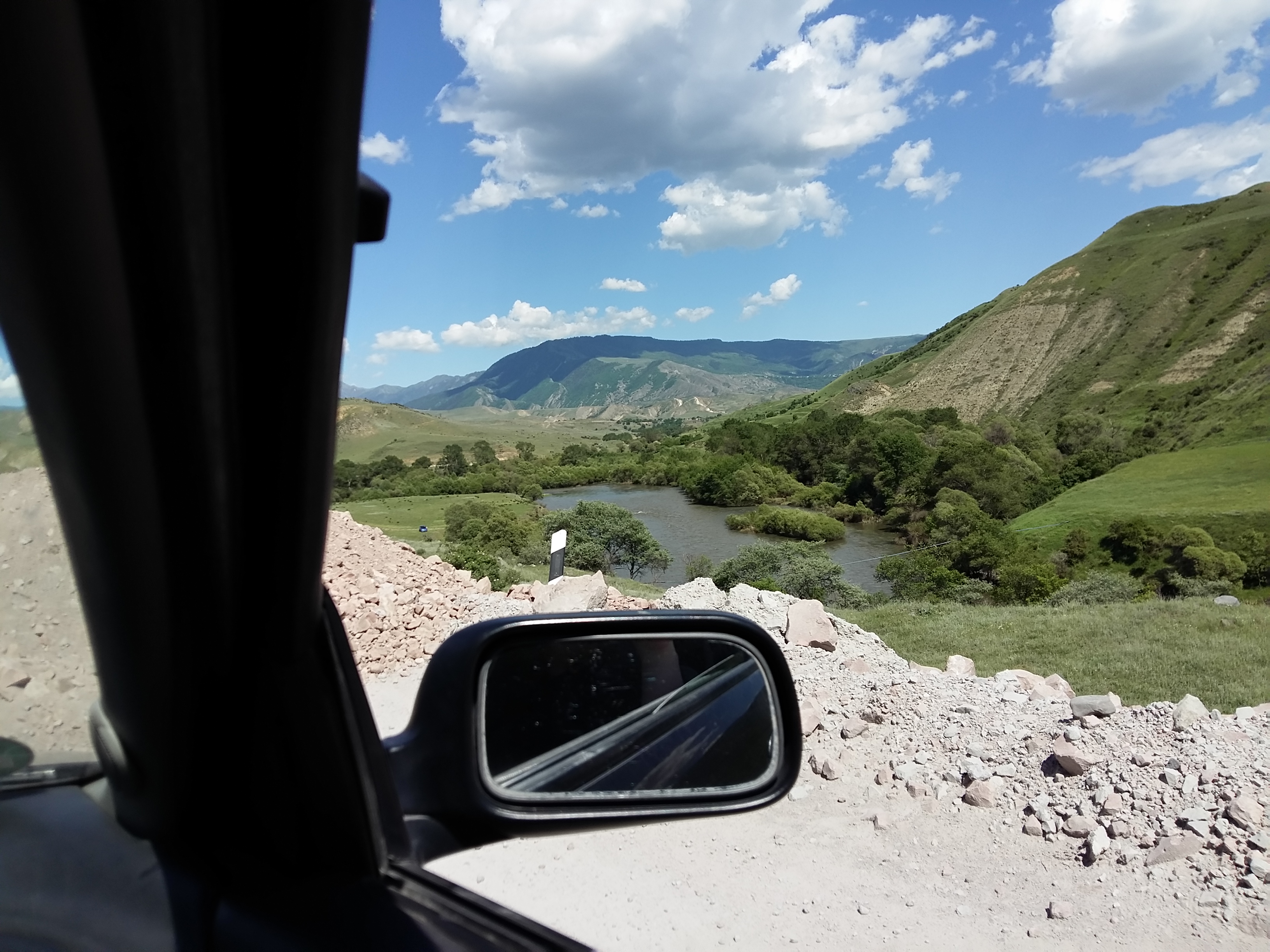 Rubble at the side of the road, seen through a car window. Behind it, a landscape with a river and mountains.