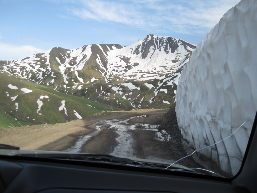 Mountain road beside melting snowpack that is taller than a car.