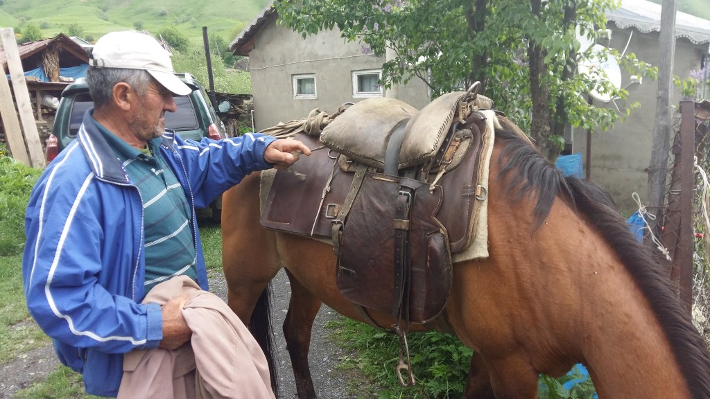 Image of a man pointing to a horse with a traditional Caucasian saddle that includes skirts and fenders from a vintage cavalry saddle.